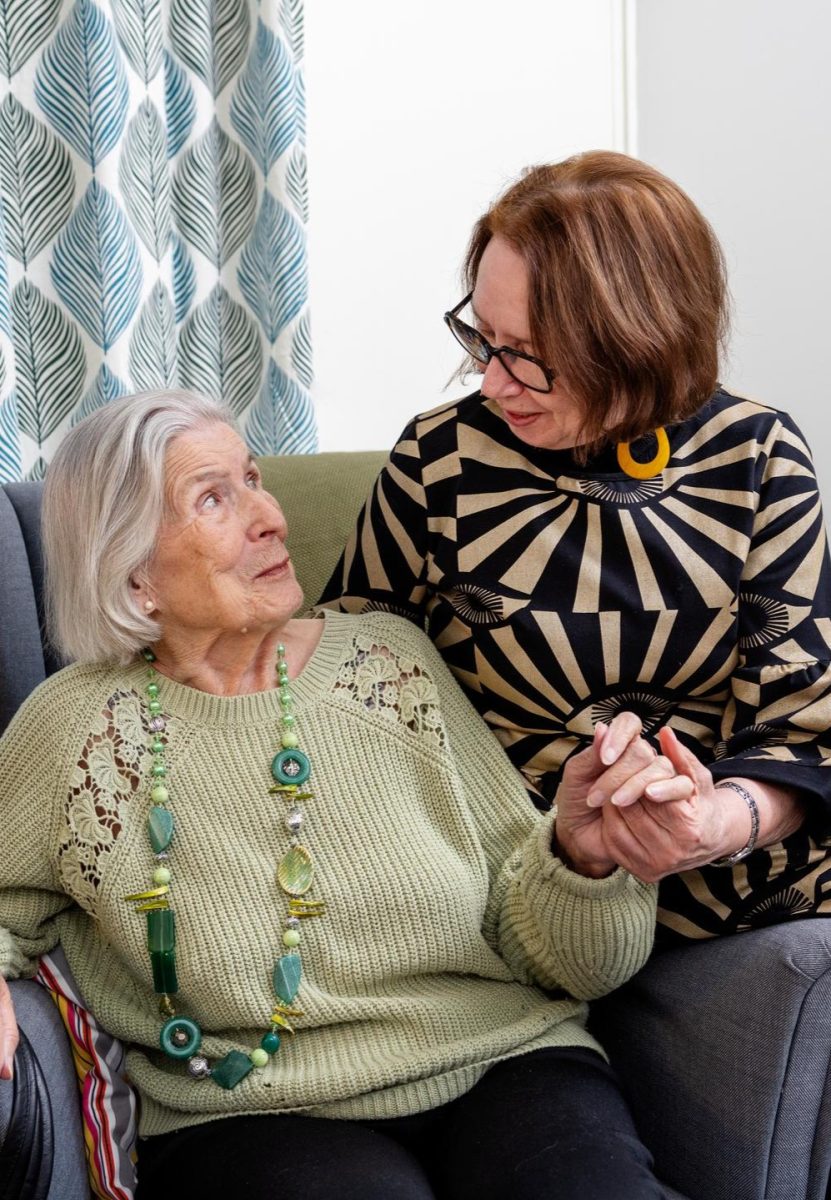 Iäkäs nainen istuu nojatuolissa. Käsinojalla istuu tukija, joka pitää naista kädestä. Tunnelma on lämmin.