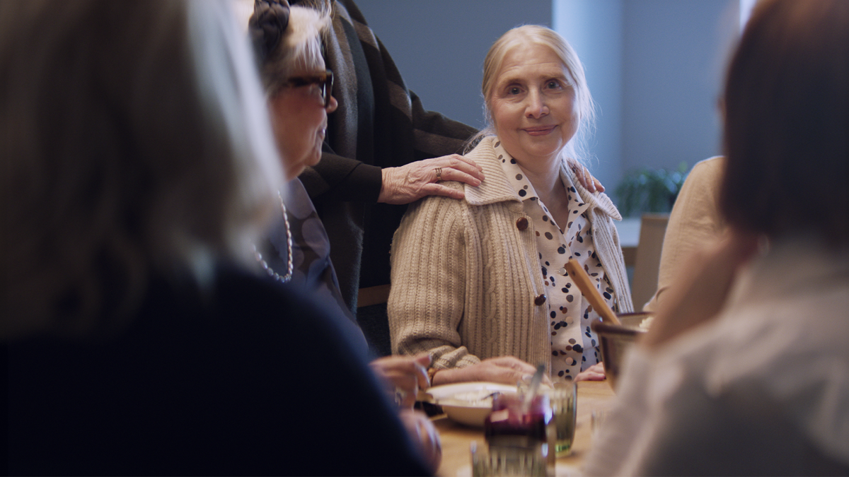 Ikäihminen on istunut pöytään, jossa on seuraa ympärillä. Yksi heistä on laskenut kädet vanhuksen hartioille.
