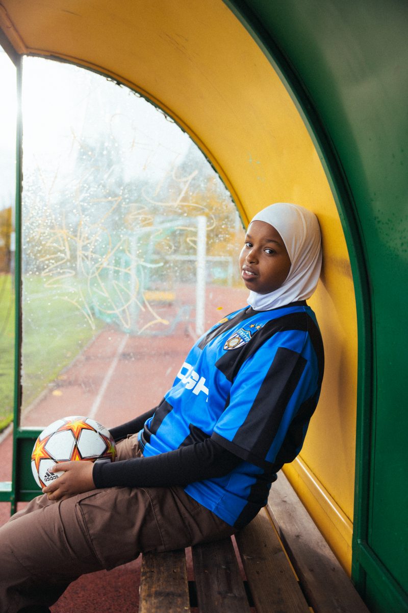 Tyttö istuu katoksessa jalkapallo sylissä ja katsoo kameraan.