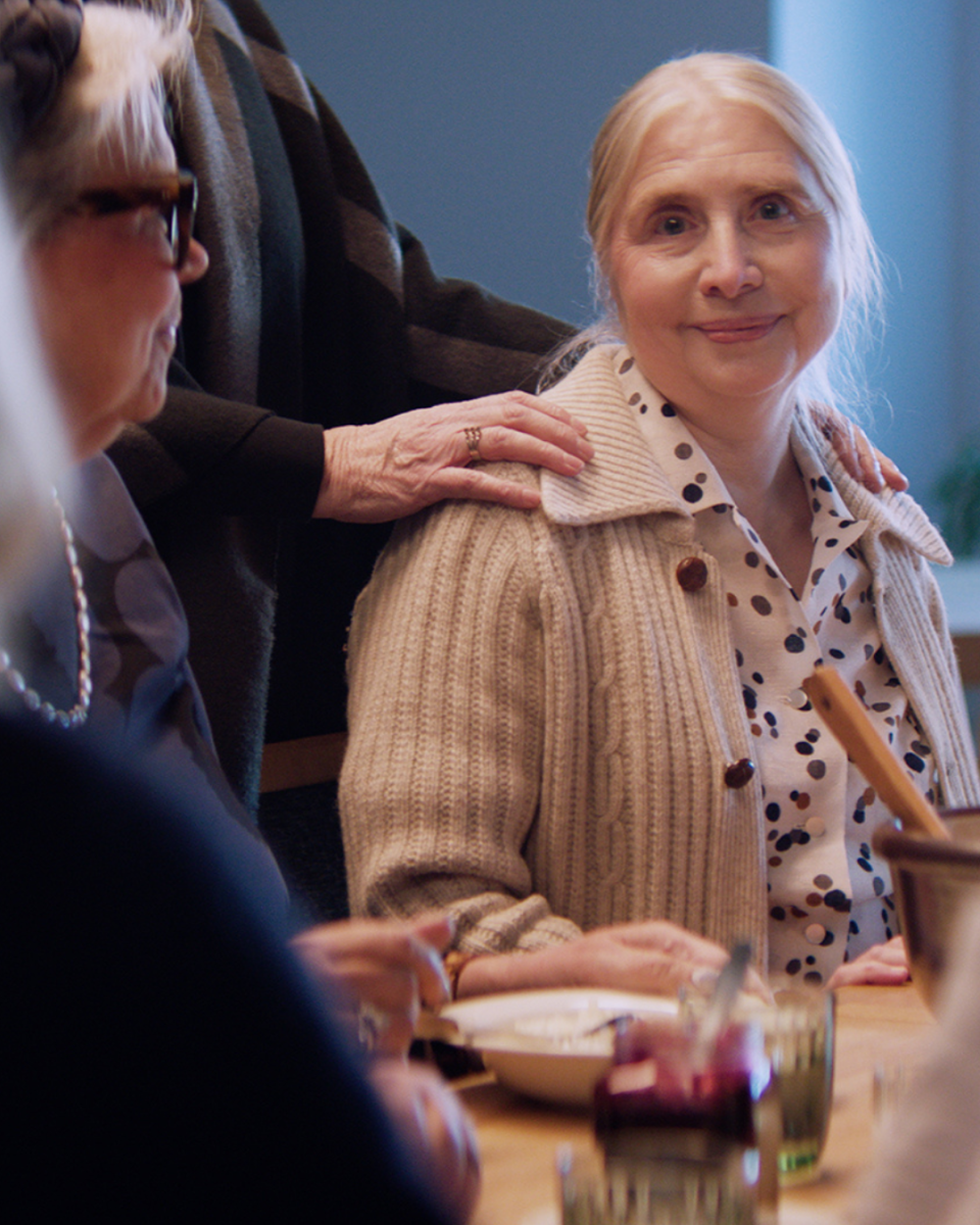 Ikääntyvä nainen on istunut pöytään, jossa on seuraa ympärillä. Yksi heistä on laskenut kädet vanhuksen hartioille.