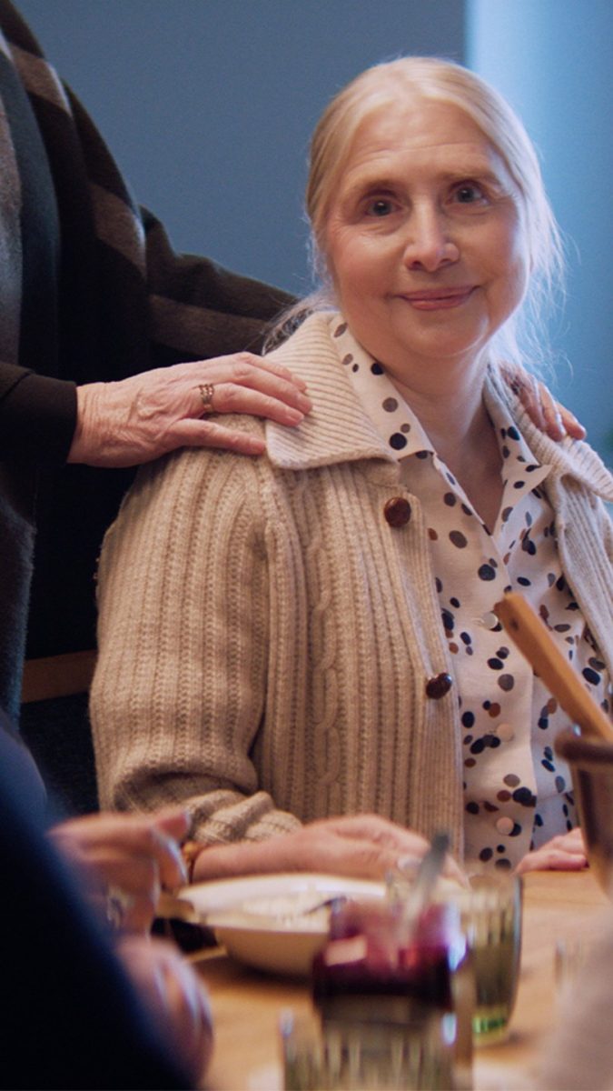 Ikääntyvä nainen on istunut pöytään, jossa on seuraa ympärillä. Yksi heistä on laskenut kädet vanhuksen hartioille.